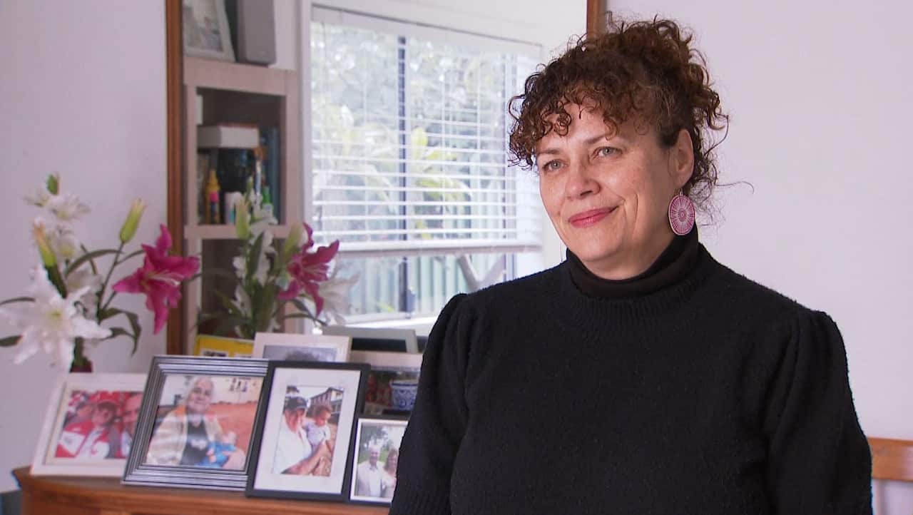 A woman with curly hair is wearing a black sweater and smiling as she sits in front of a mirror and a console table decorated with family photographs. 