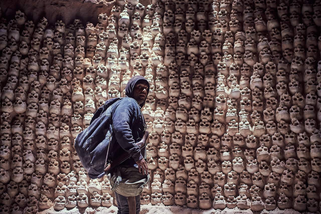 A man stands in a hidden cave full of stacked human skulls.jpeg