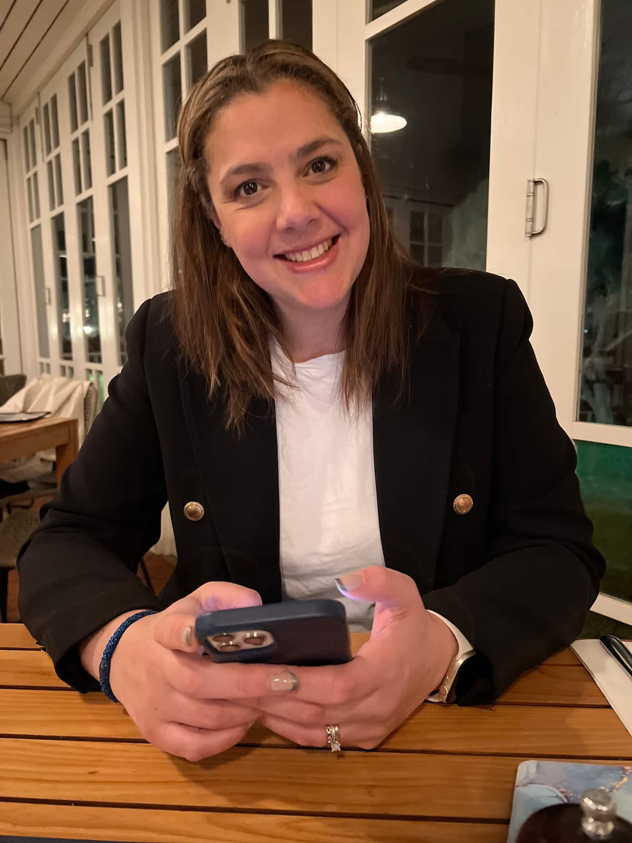 A picture of a woman smiling as she sits at a table and holds her mobile phone.