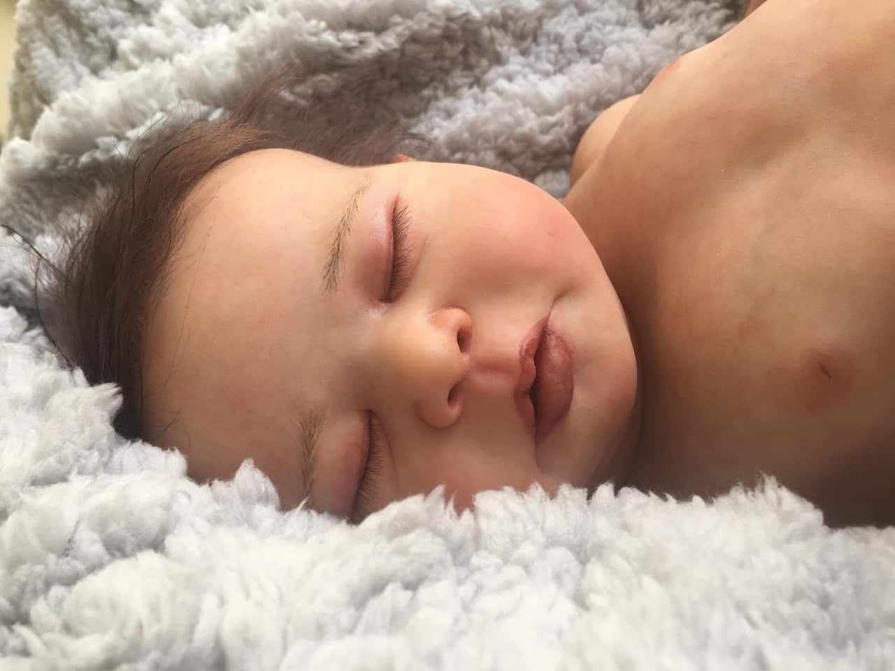 A doll resembling a sleeping human infant lies on its side on a fluffy blanket 