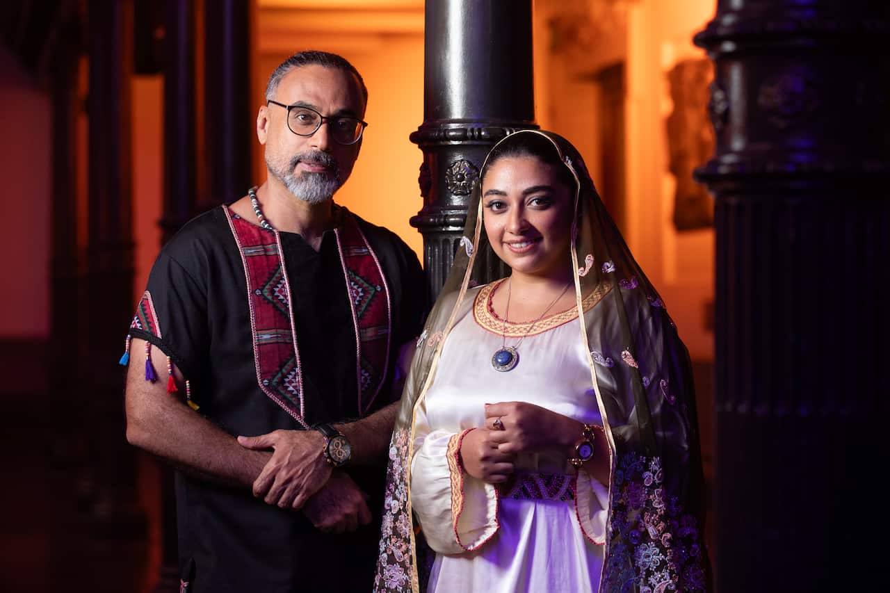 A man and woman wearing traditional clothing stand beside a pillar, smiling at the camera.