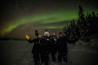 Four Indigenous rangers wearing puffer jackets, standing in the snow at night, with an aurora in the background.