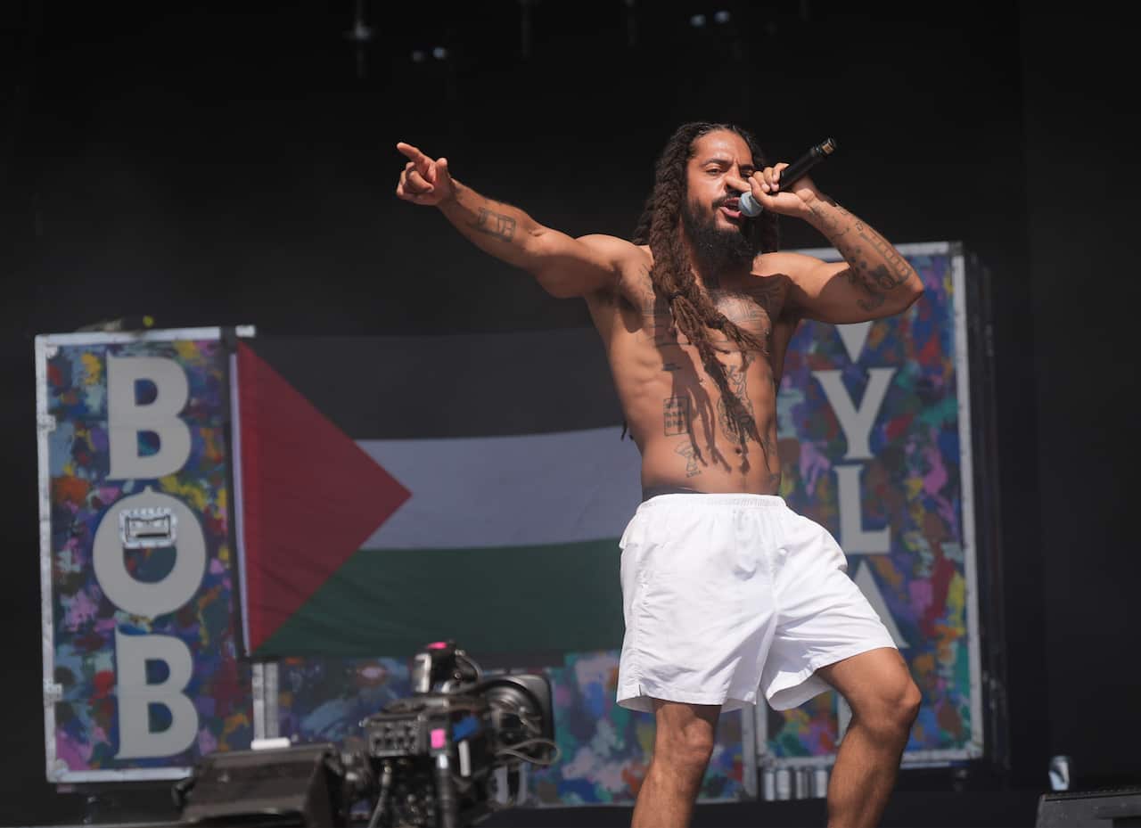 A man on stage rapping in front of a sign that says "BOB VYLAN" with the Palestinian flag.