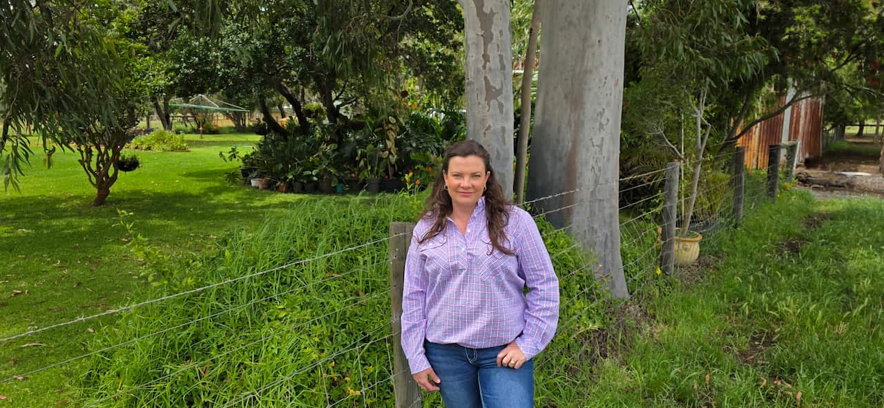 A woman in a checked shirt and jeans stands in a field next to a wire fence, with a light smile on her face.