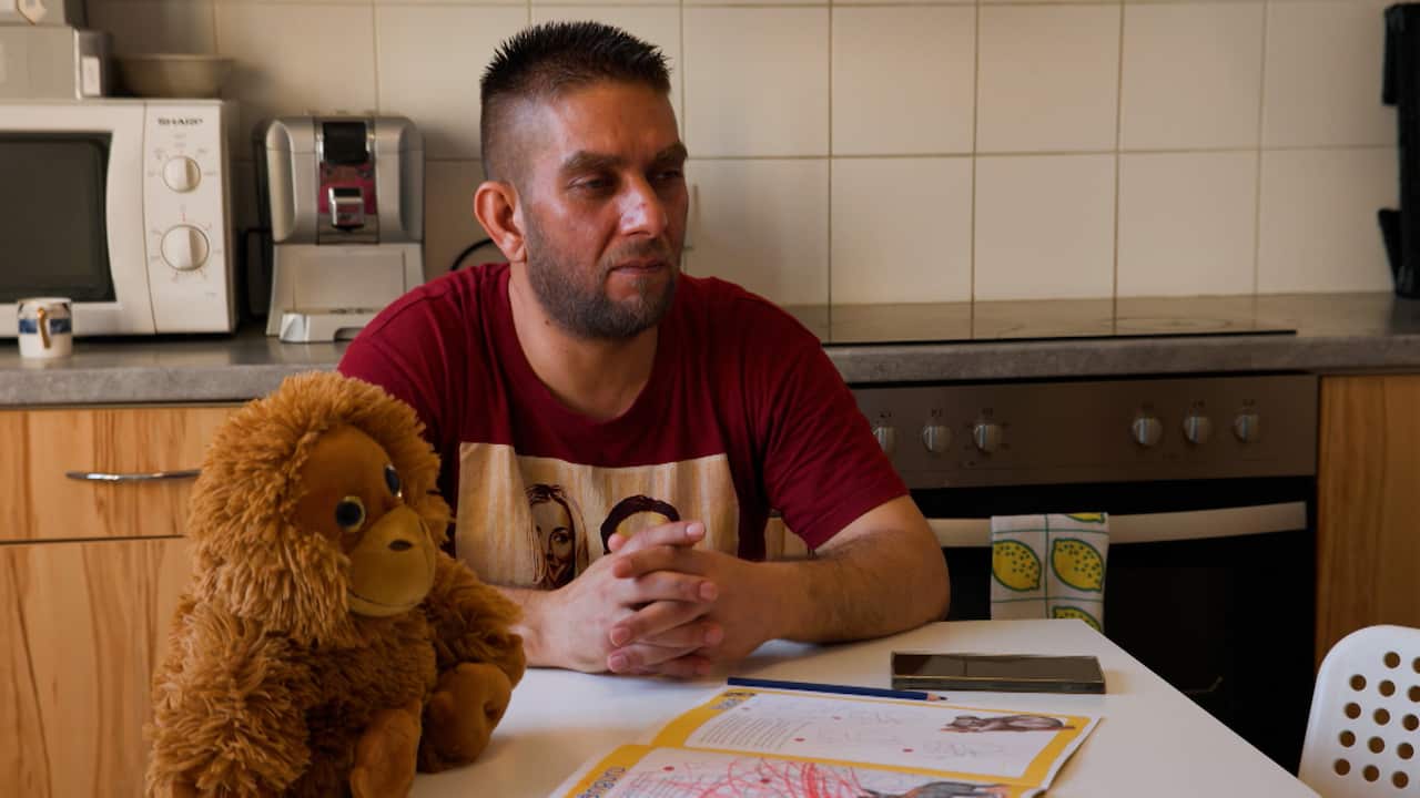A man sits at a kitchen table with his hands clasped, looking thoughtfully to the side next to a plush orangutan toy and an open coloring book.