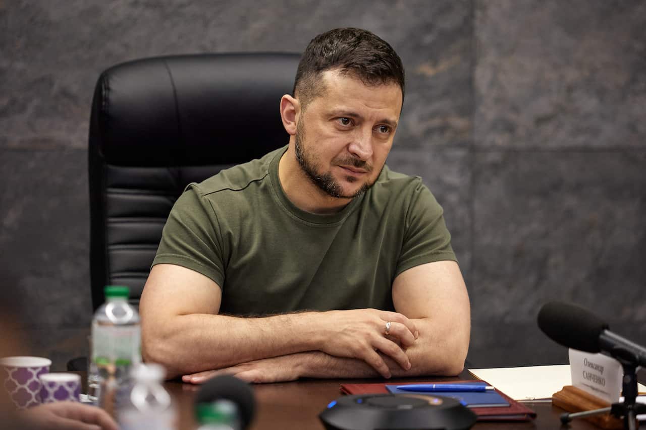 Volodymyr Zelenskyy sits at a desk wih his arms overlapped. 