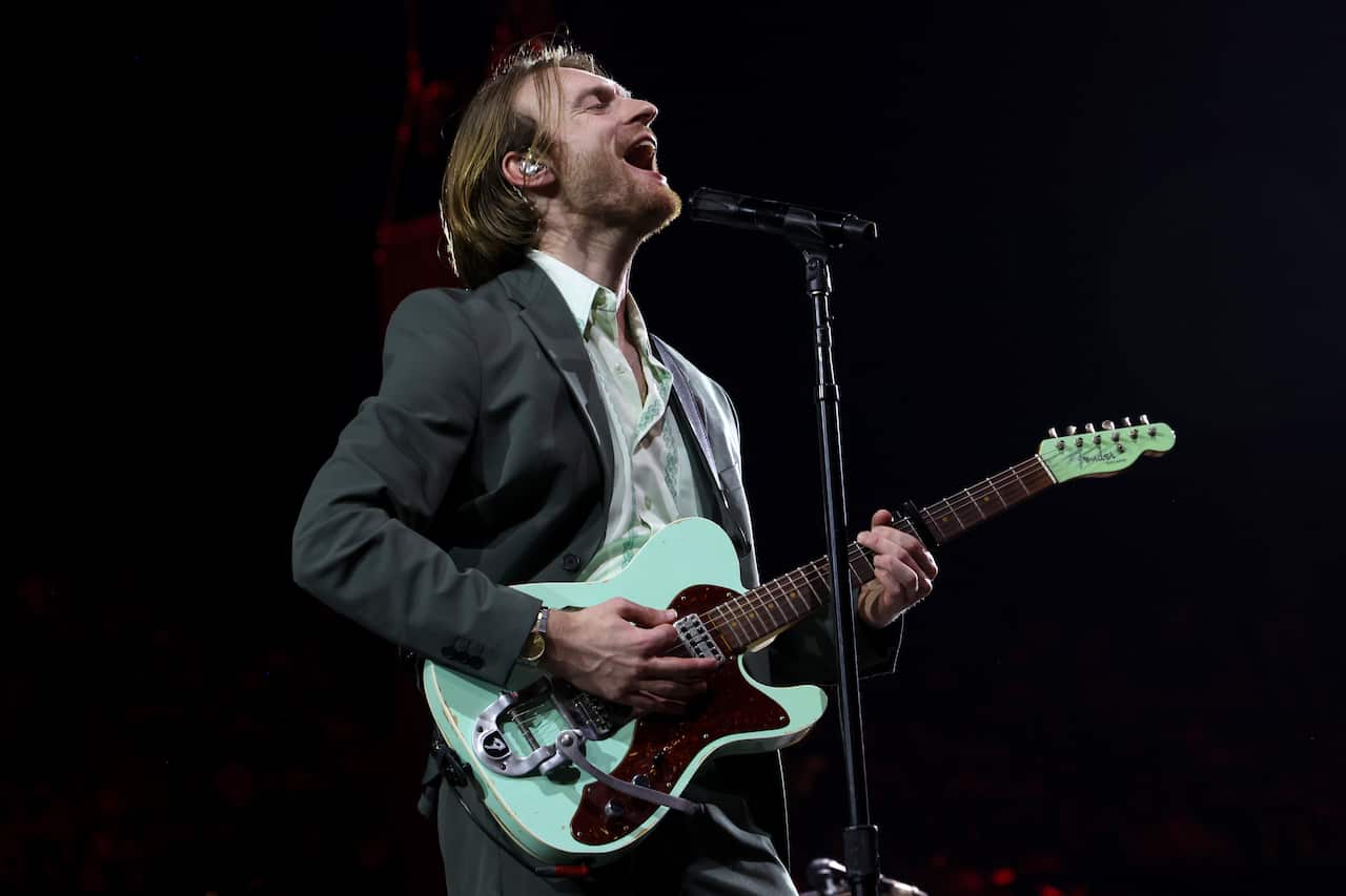 Finneas, wearing a white shirt and grey suit jacket, playing a mint-coloured electric guitar and singing into a microphone on a stand