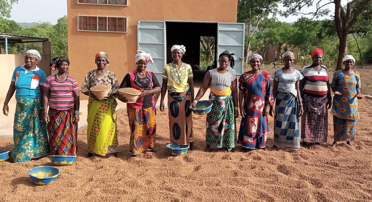 African women dressed in bright colours stand in a line on red sand in front of a building