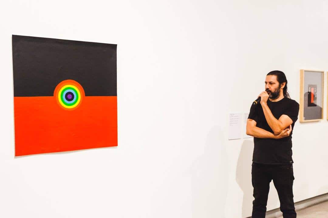 Archie Moore in black jeans and t shirt stands in front of one of his artworks with microphone at an exhibition in 2017