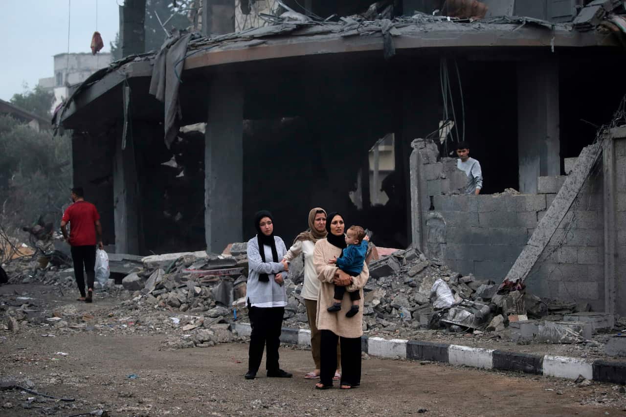 Women and children stand in front of a destroyed building. 