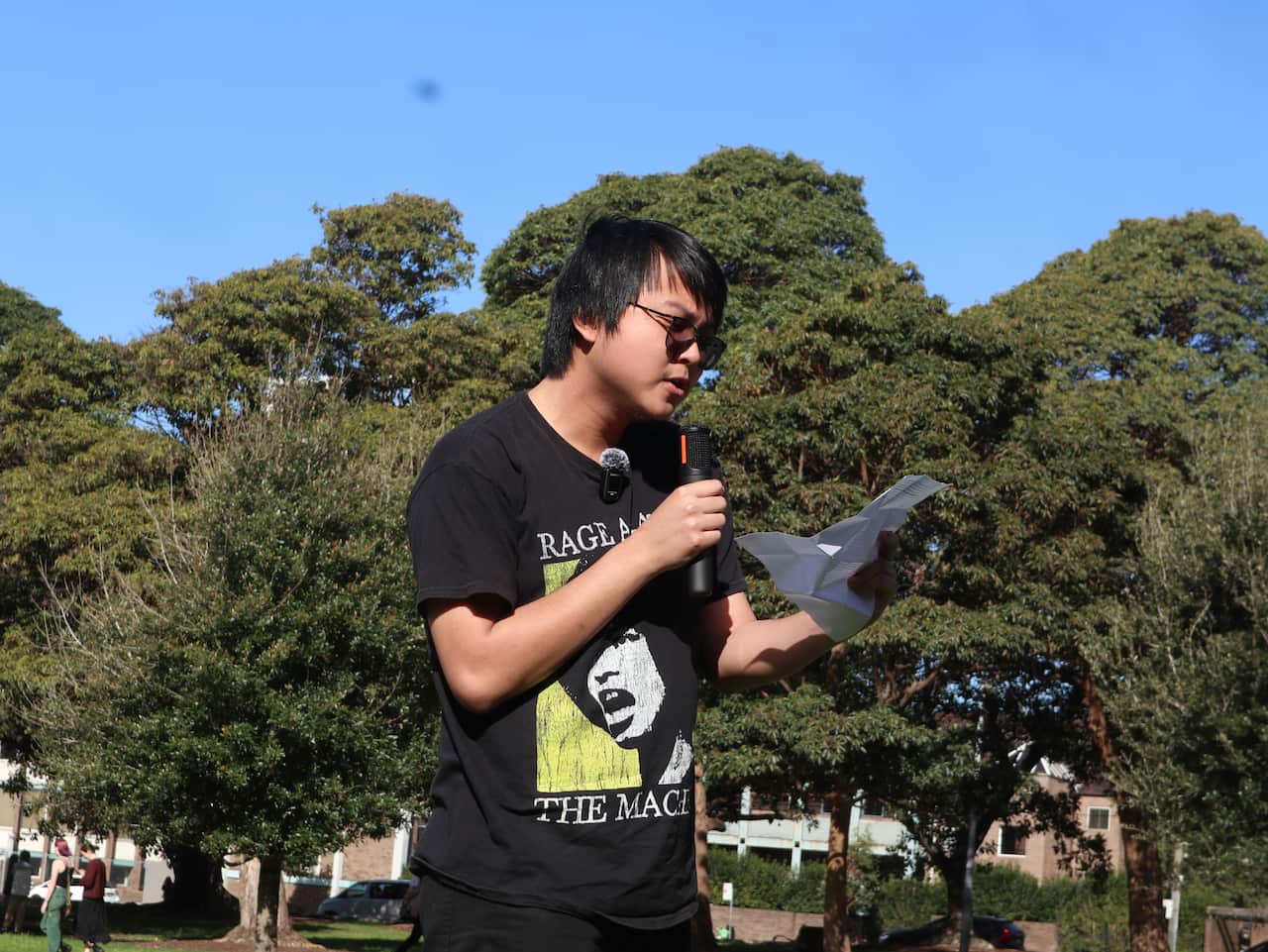 A man in a black round-neck t-shirt is reading from a sheet of paper as he speaks into a microphone.