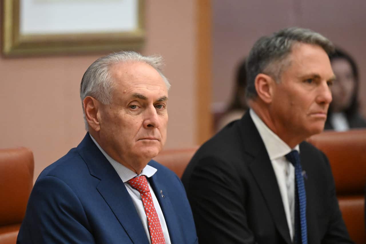 Two older white men in suits sit beside each other looking serious.