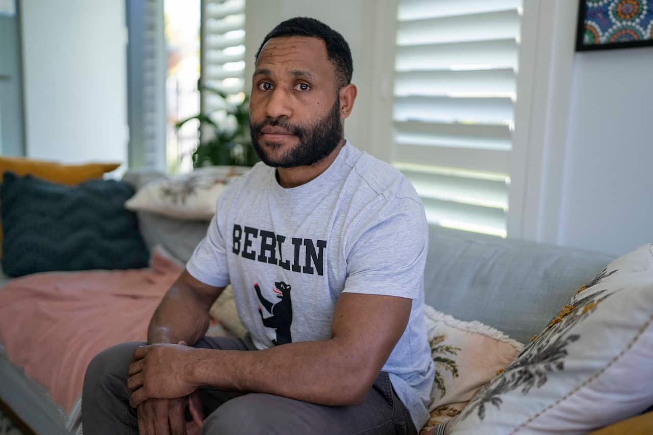 A PNG man wearing a grey marle t-shirt with the word Berlin written on it and an image of a bear below is sitting on a couch in a living room.
