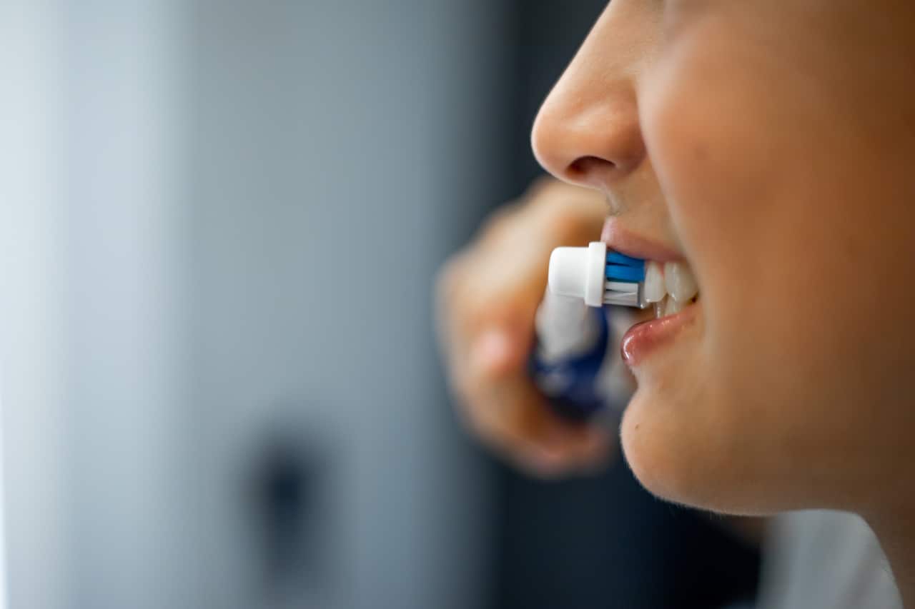 Close up mouth of teenage boy holding electric toothbrush in hand, smiling and brushing teeth in bathroom.