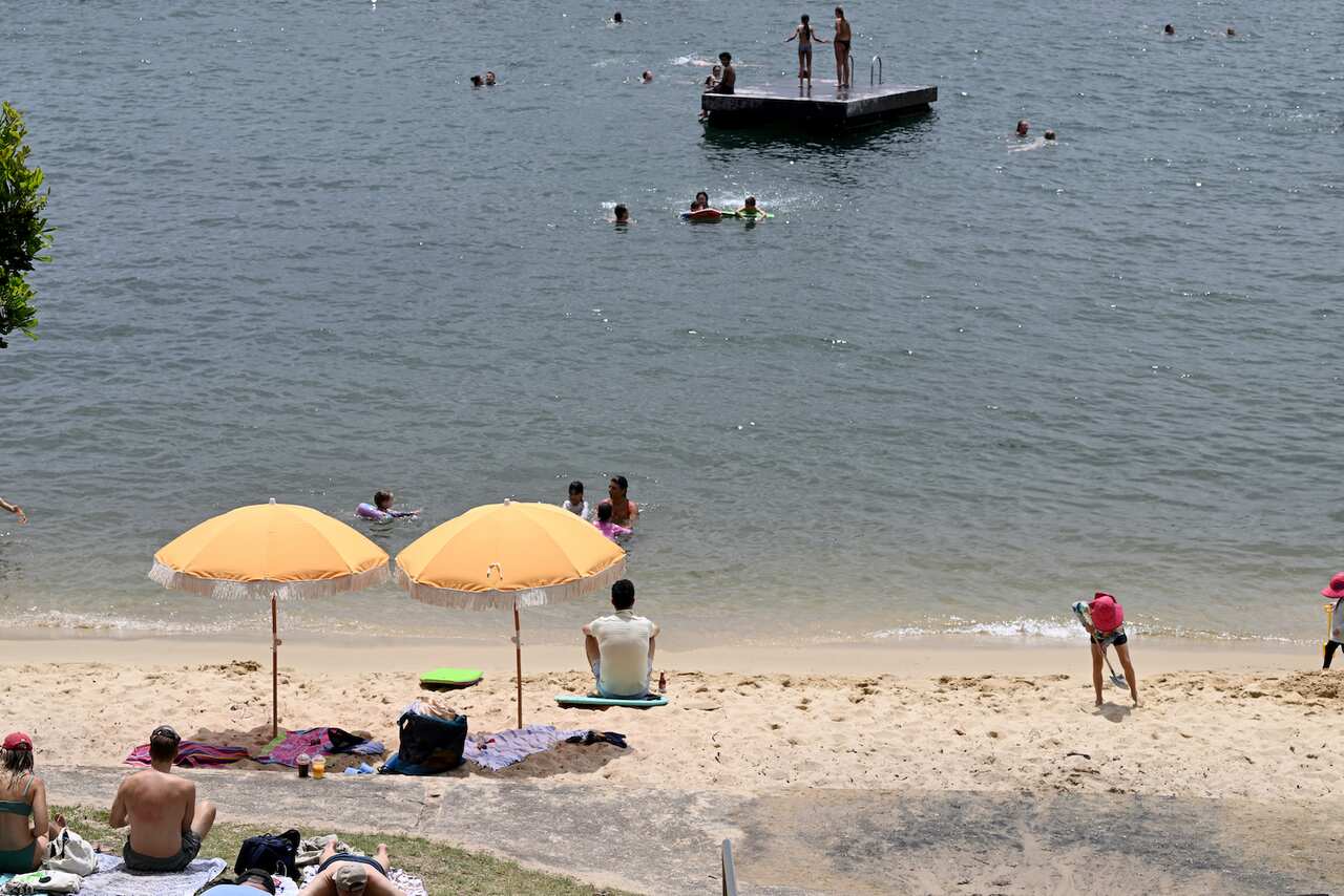 A beach with sand, parasols and people sitting. The water is visible, with people swimming in it.