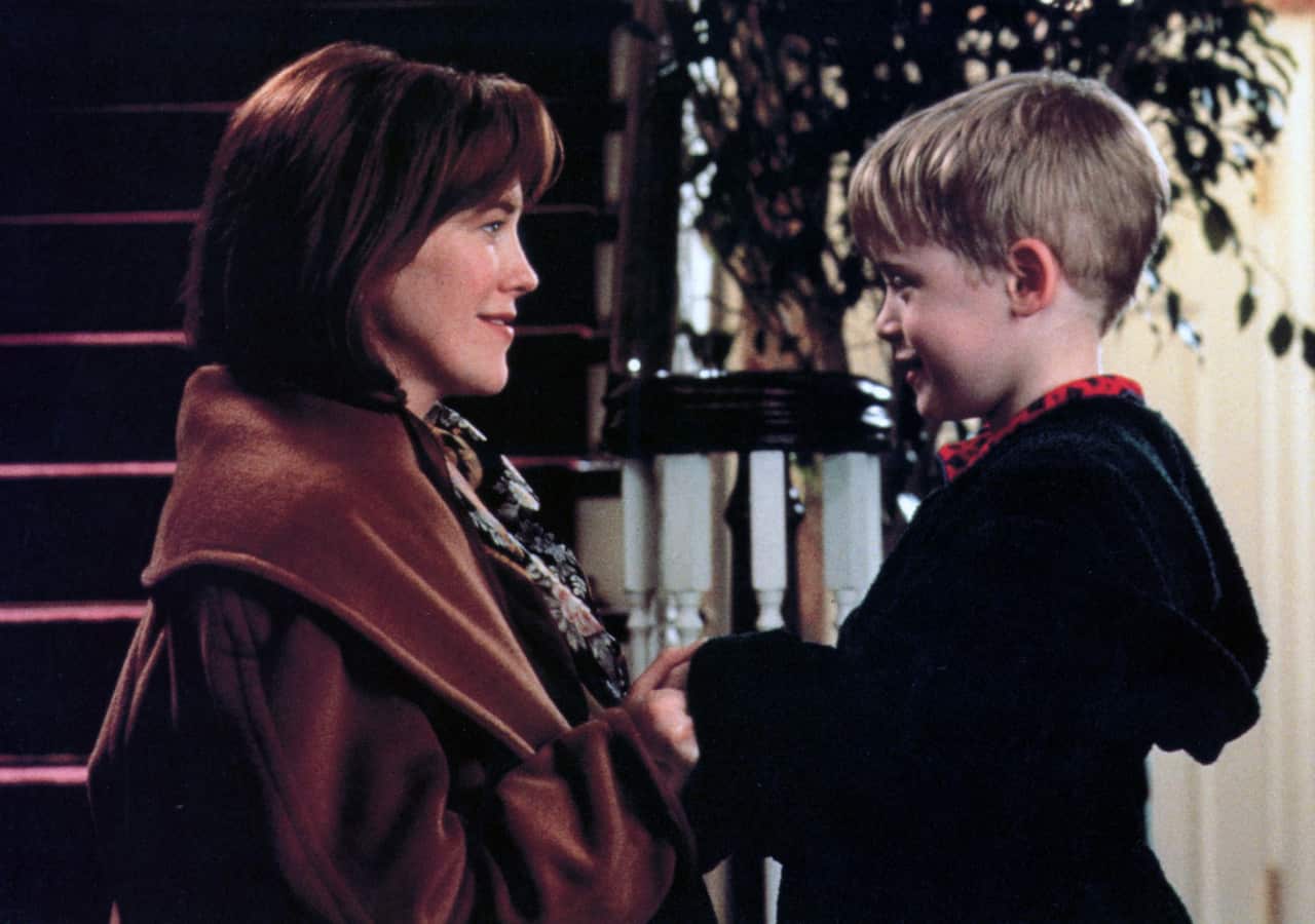 A woman and a young boy face each other indoors near a staircase, holding hands and smiling