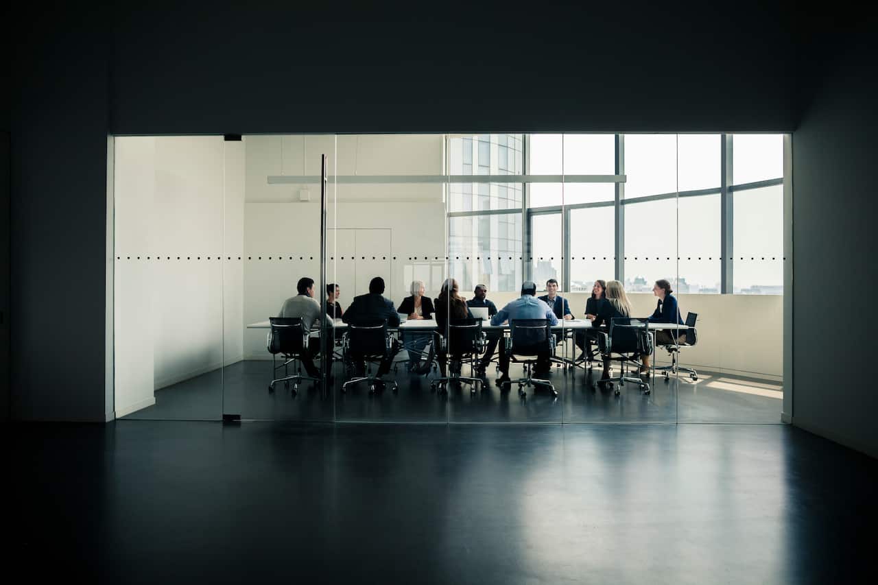 A group of people sitting on chairs around a table in a closed off room with glass doors.