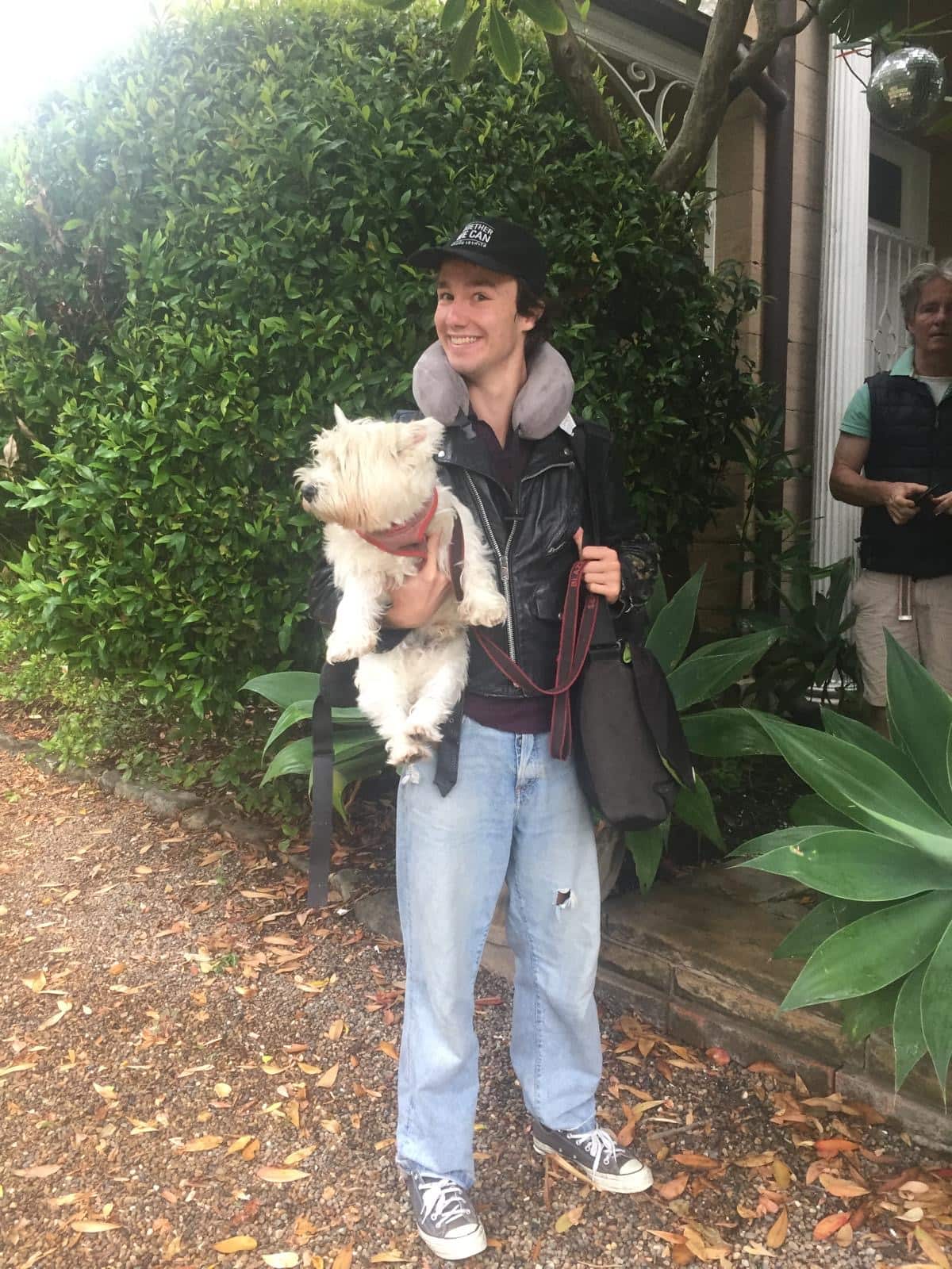 A man wearing a black cap and a black jacket with blue jeans standing in front of a green bush holding a white dog.