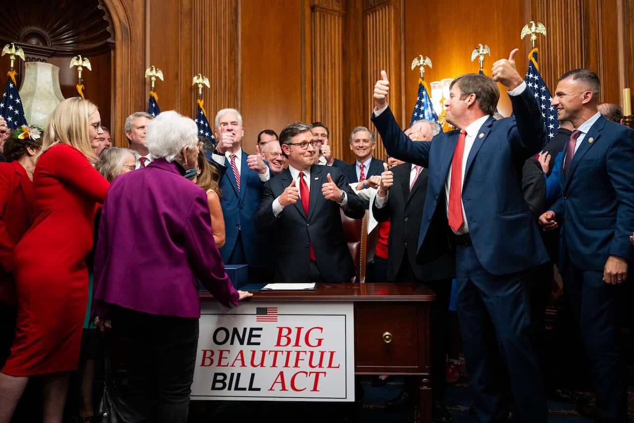 A large group of politicians cheers and raises their thumbs. A sign that says "One Big Beautiful Bill Act" is attached to a desk.