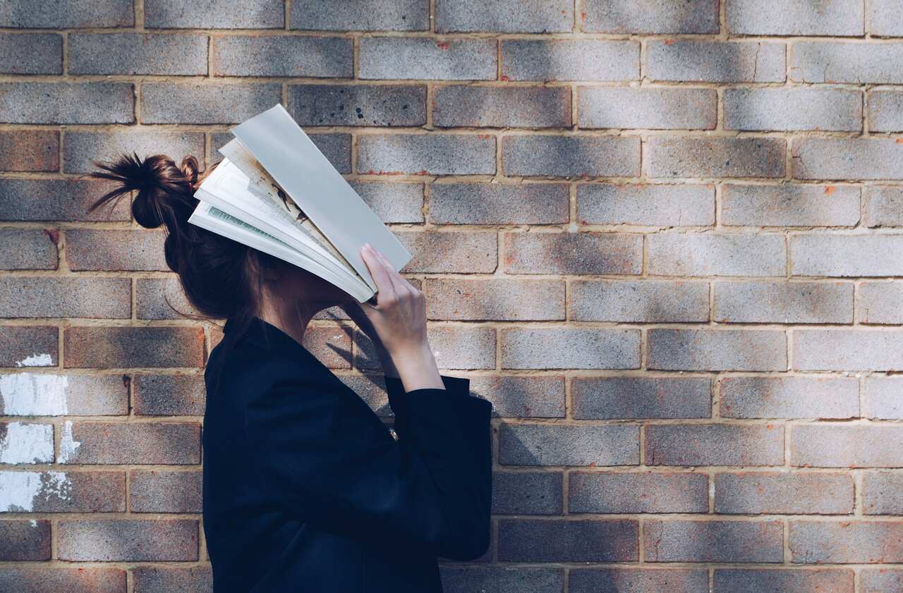School Girl with Book in front of natural rustic red brick background holding book up to her face. Picture courtesy of Siora Photography 