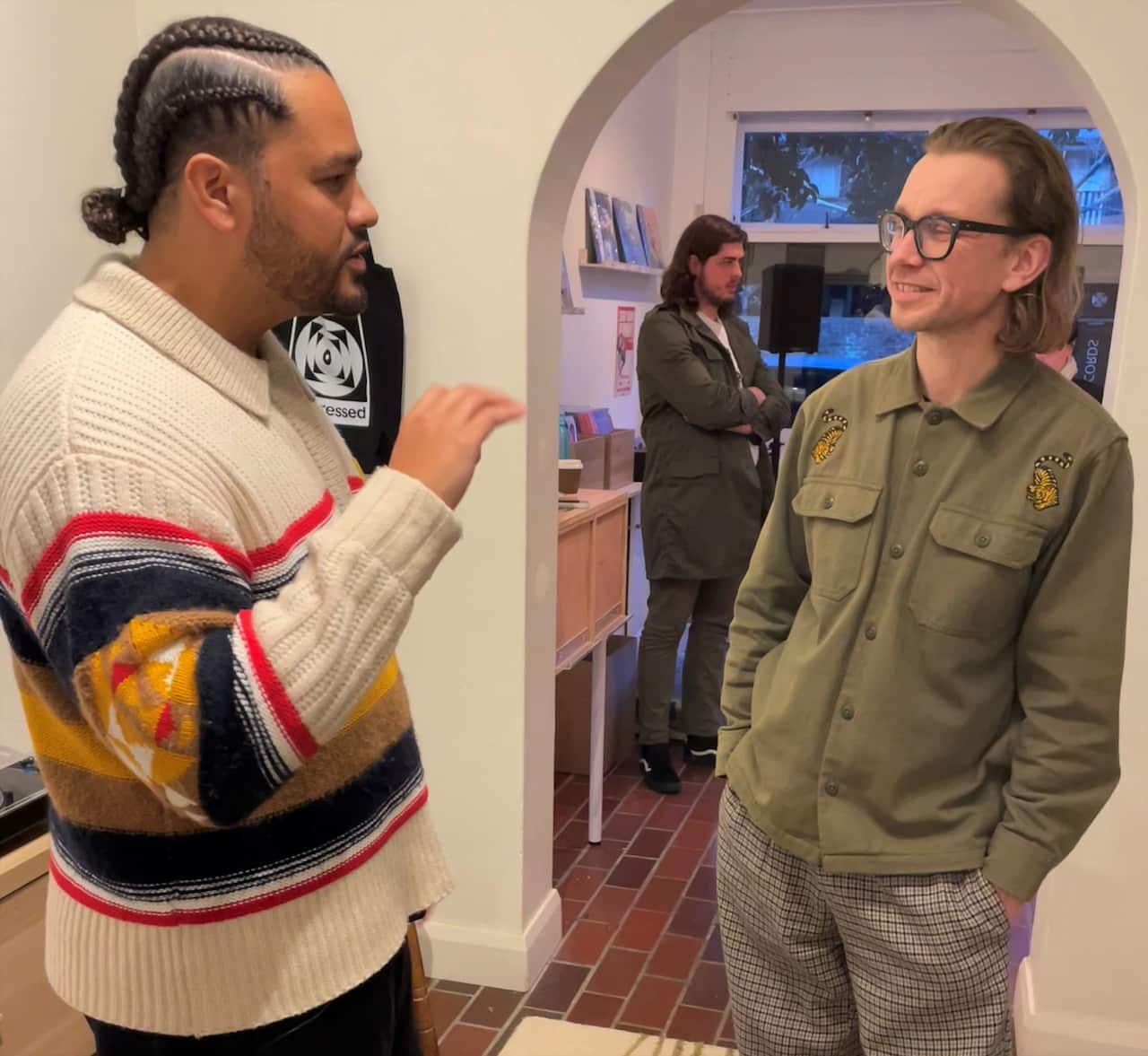 A man in a jumper stands talking to another man in a khaki jacket.