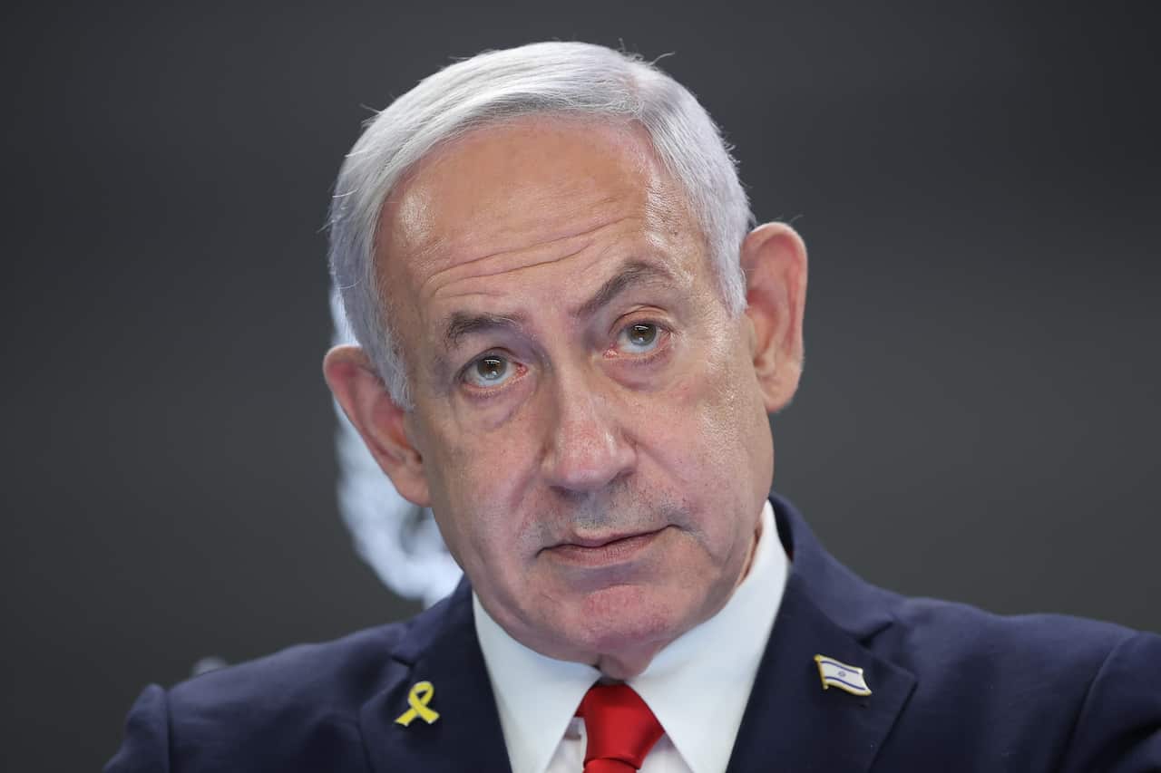 Benjamin Netanyahu, wearing a dark blue suit, whit shirt and red tie. There is an Israeli flag pin on his jacket.