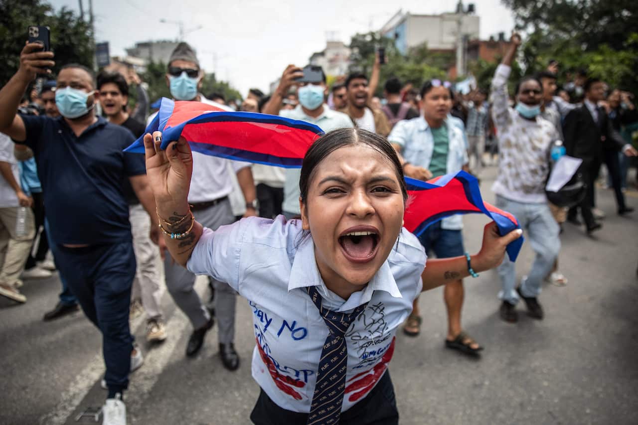 A young girl holding a flag screams, with protesters walking and shouting slogans behind her during a rally.
