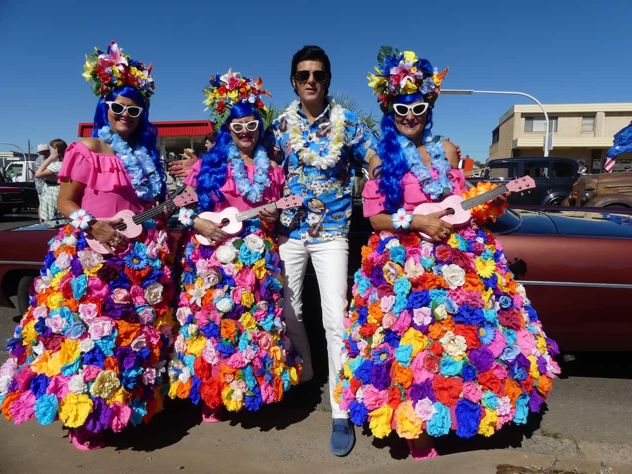 A man wearing dark sunglasses, a lei, a floral shit and white pants. Three women with blue hair, wearing floral dresses and holding ukuleles stand next to him.