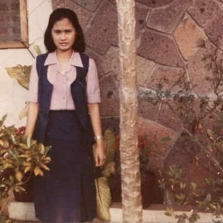 A young woman stands outside a home. She is looking at the camera with a neutral expression on her face.