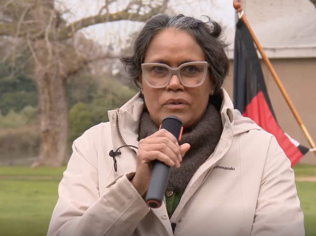 Cathy Freeman speaking into a microphone while standing in front of an Indigenous flag.