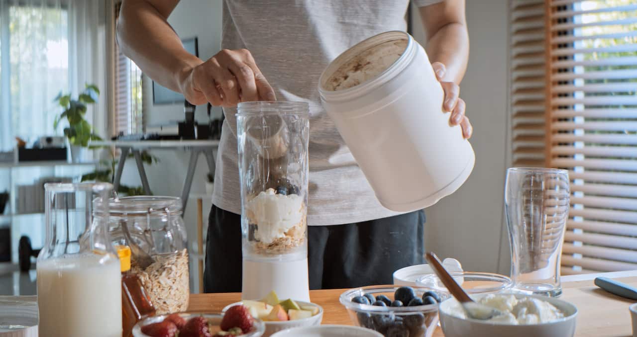 A person holding a tub of powder in their left hand. In their right hand they are adding a scoop of powder to a long clear container with other food ingredients in it.