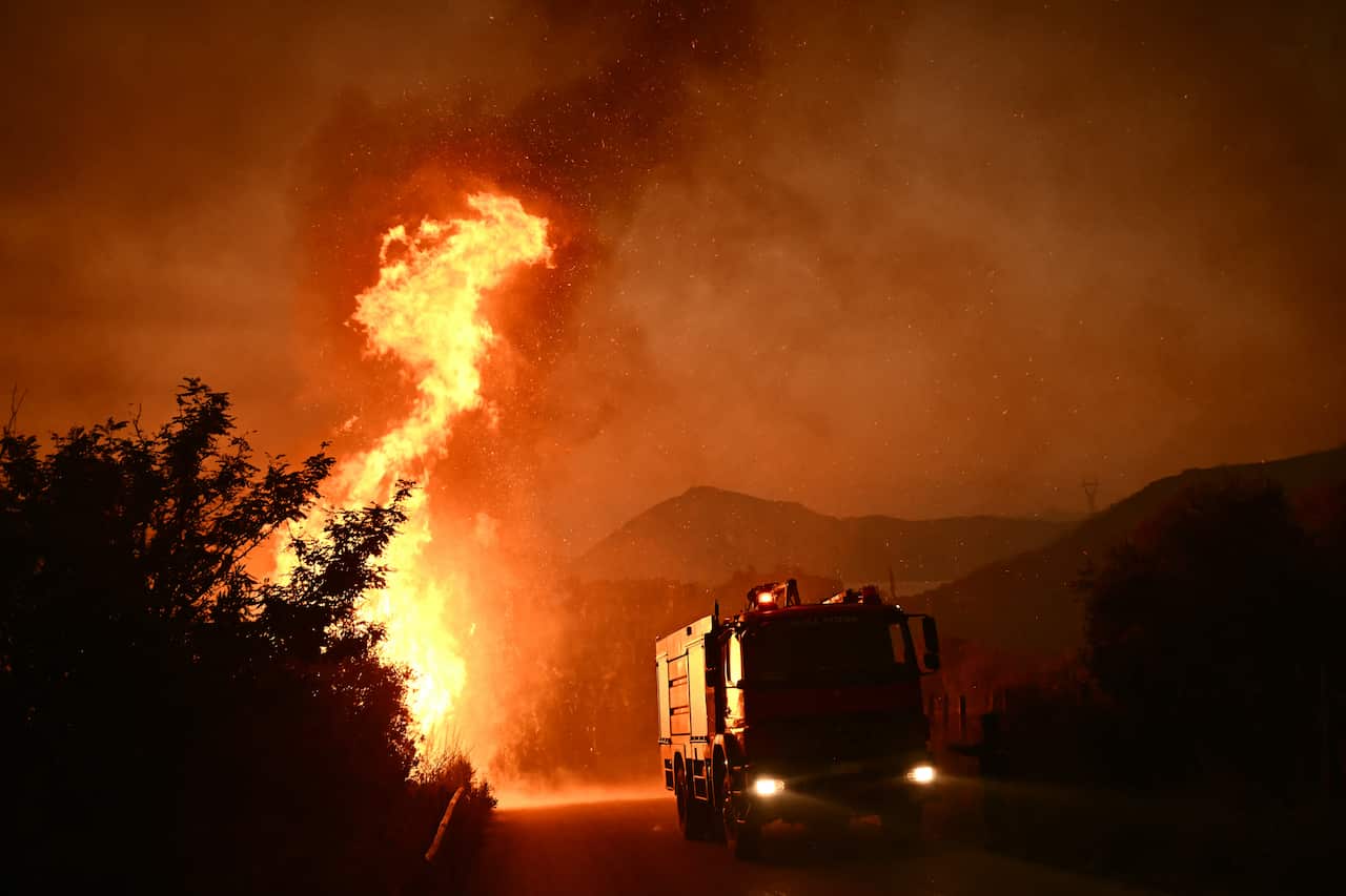 A firetruck drives past flames along a road during a wildfire.
