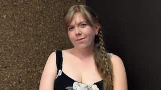 A close-up of a young woman with dark blonde hair in a plait wearing a black, floral-patterned dress