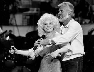 Two people, a younger woman and an older man, dance while holding microphones and smiling.
