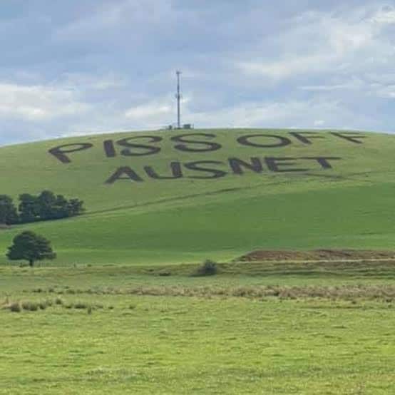 A grassy green hill with the words PISS OFF AUSNET mowed into the grass in large capital letters. A single communication tower stands at the top of the hill. 
