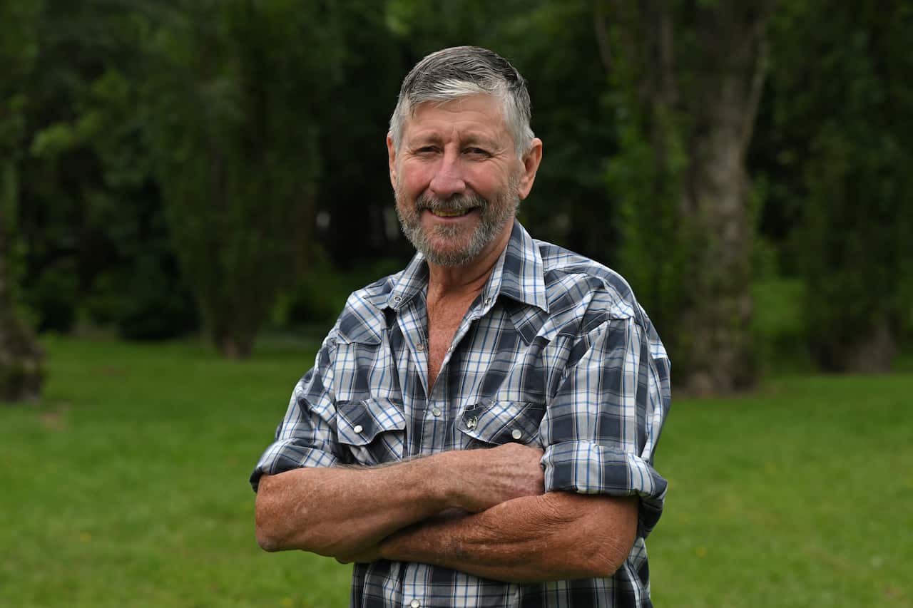 A grey-haired man wearing a chequered shirt stands with his arms crossed