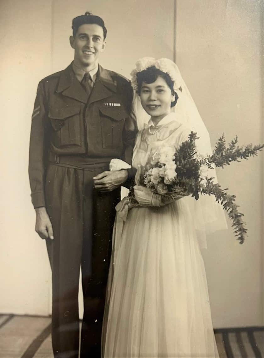 An old photo of an Australian soldier and a Japanese woman in a white dress, holding a bouquet, taken on their wedding day.