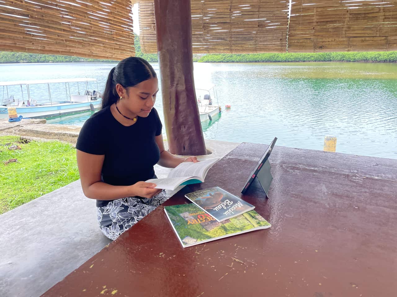 A young girl wearing a black top and printed bottom sits at a wooden table next to a body of water, reading a book.
