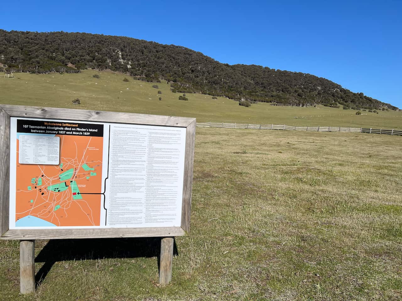 A sign in an open area of land. There is a tree-covered hill in the background