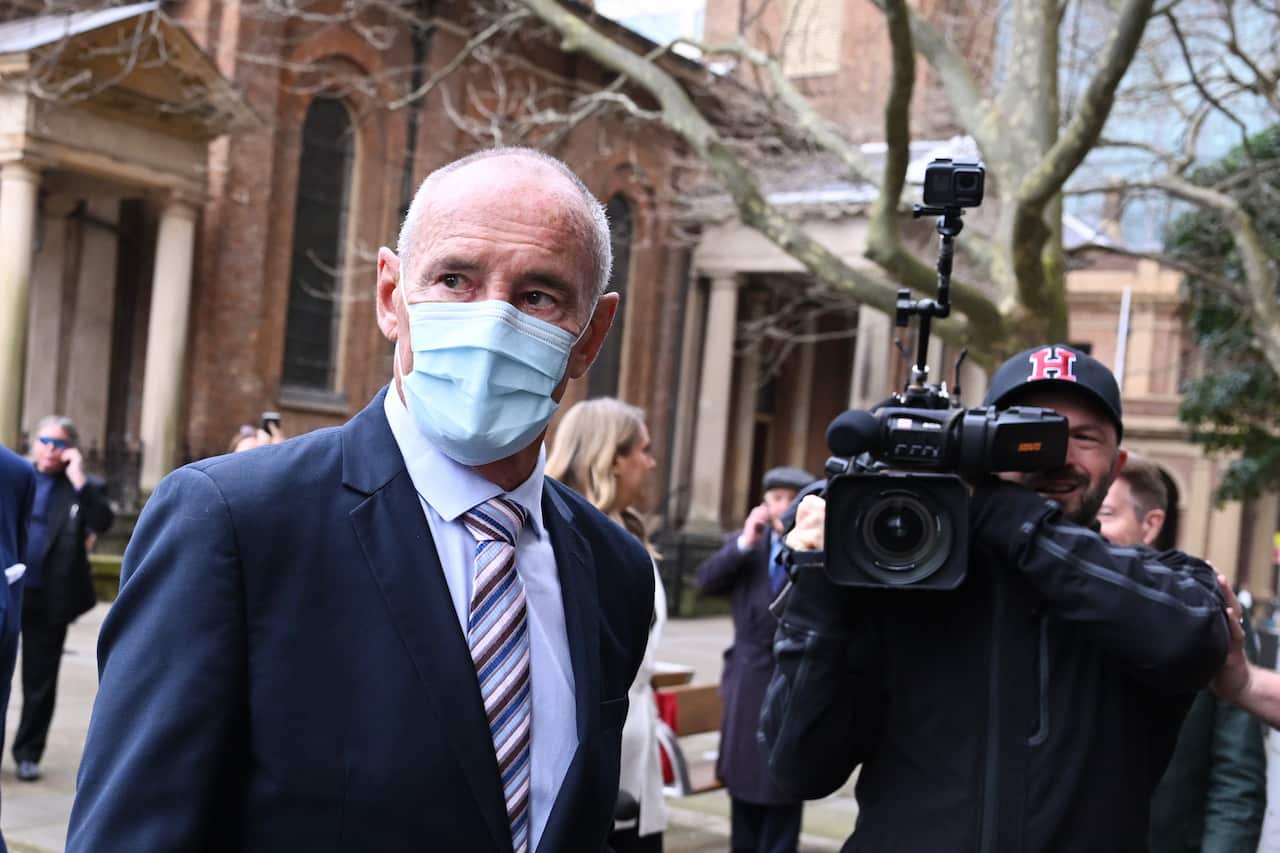 A man wearing a face mask stands outside the NSW Supreme Court, while a member of the media takes his photo.