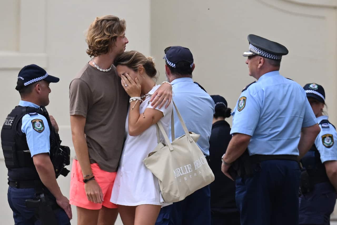 A man comforting a woman, while several police officers stand near them.