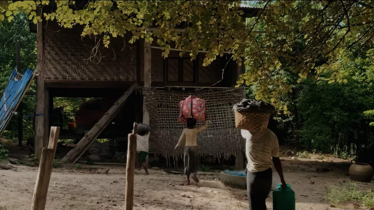 Three people carry baskets and household items as they walk toward a traditional raised bamboo house surrounded by trees.