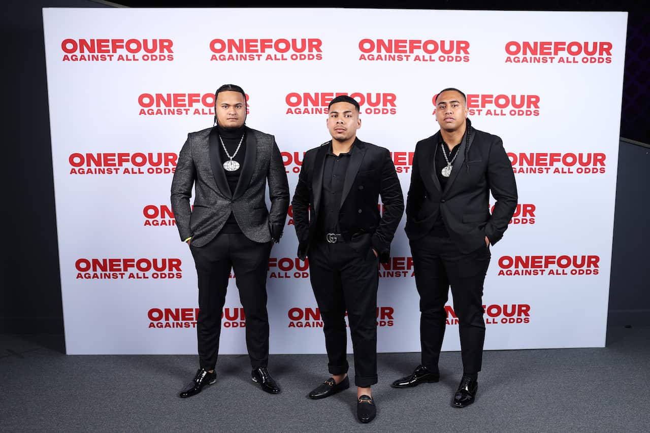Three young men dressed in formal attire at a documentary premier. They are standing in front of a screen with the documentary title on it