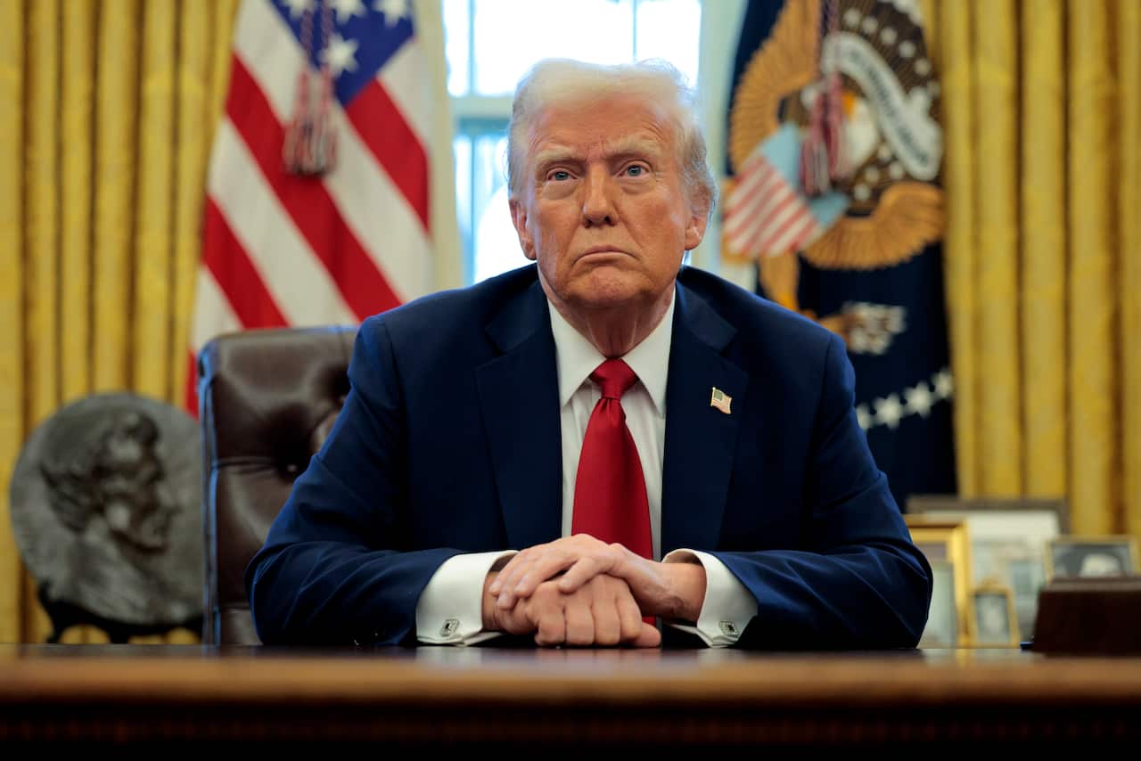 An older white man with blonde hair seated at a desk in a suit 