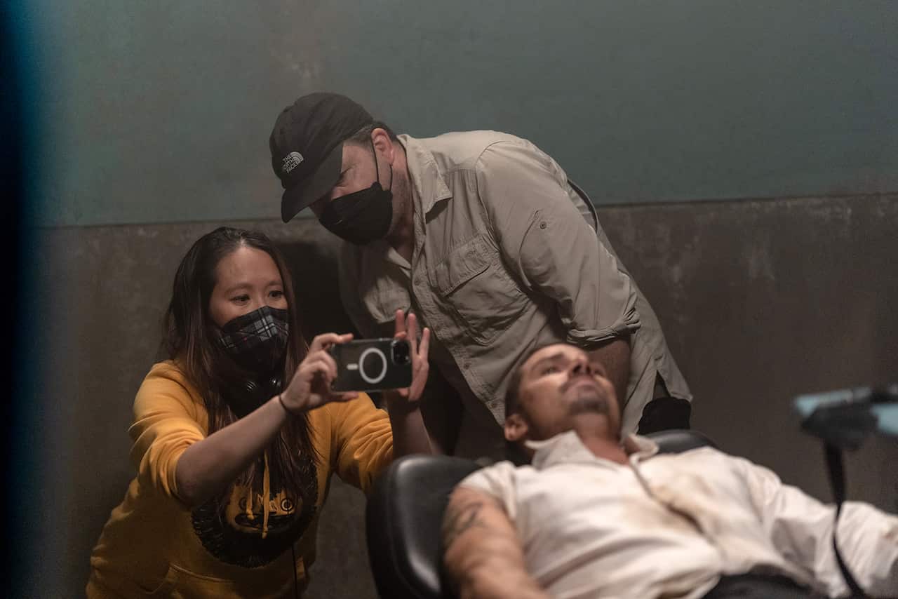A behind the scenes shot: A woman in a safety mask holds a mobile phone in front of her, aimed at a man who can be partly seen lying in a chair. Beside the woman, a man in a mask bends forward to look at the mobile phone. 