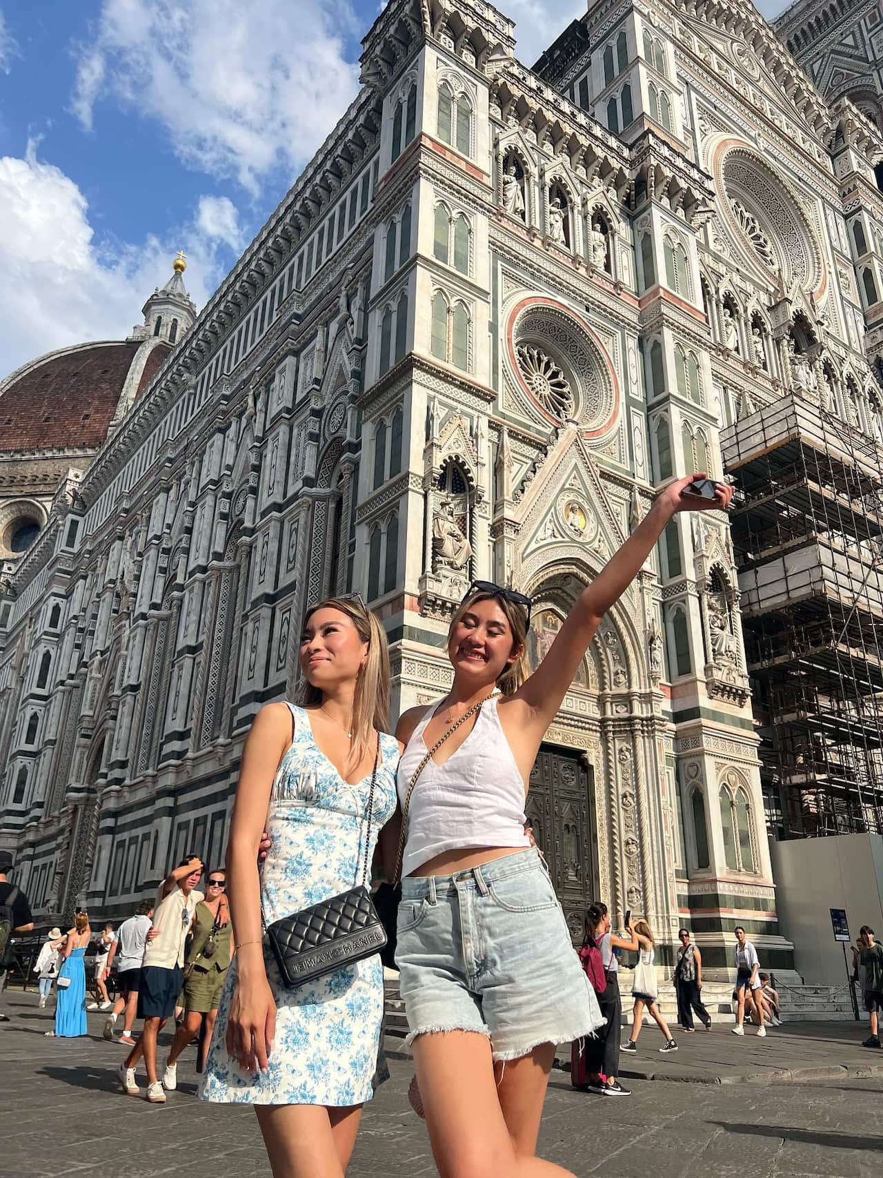 Two smiling women standing with their arms around each other, one with her other arm outstretched to the sky, in front of a church. 