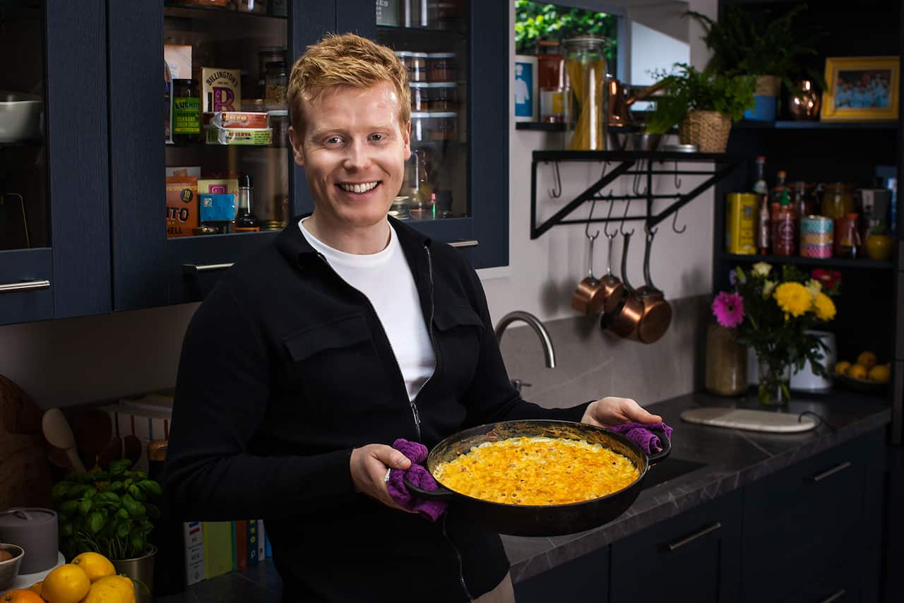 A smiling man stands in a kitchen, holding a baking dish full of a pasta bake. 
