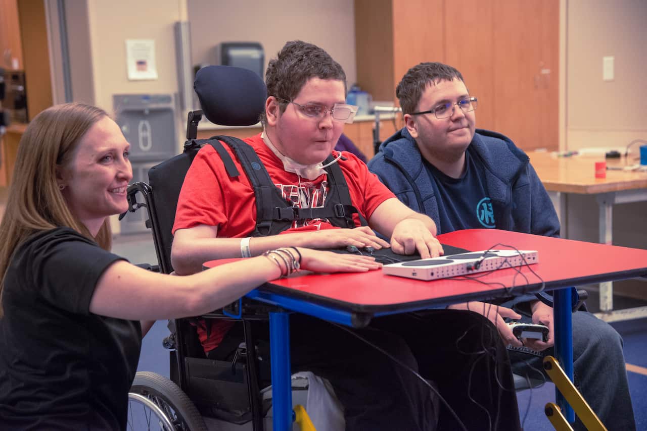A young man in a wheelchair using an adaptive video game controller placed on a red desk. There is a woman kneeling to his right and  another young man sitting to his left.