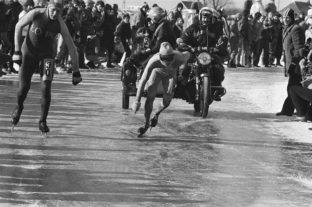 A black and white image of an ice skater racing with a competitor next to him, an old fashioned sidecar behind him and a crowd of spectators in the background