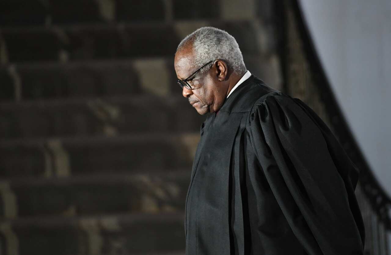 Man in black court robes with white collar and glasses has his head tilted down. 